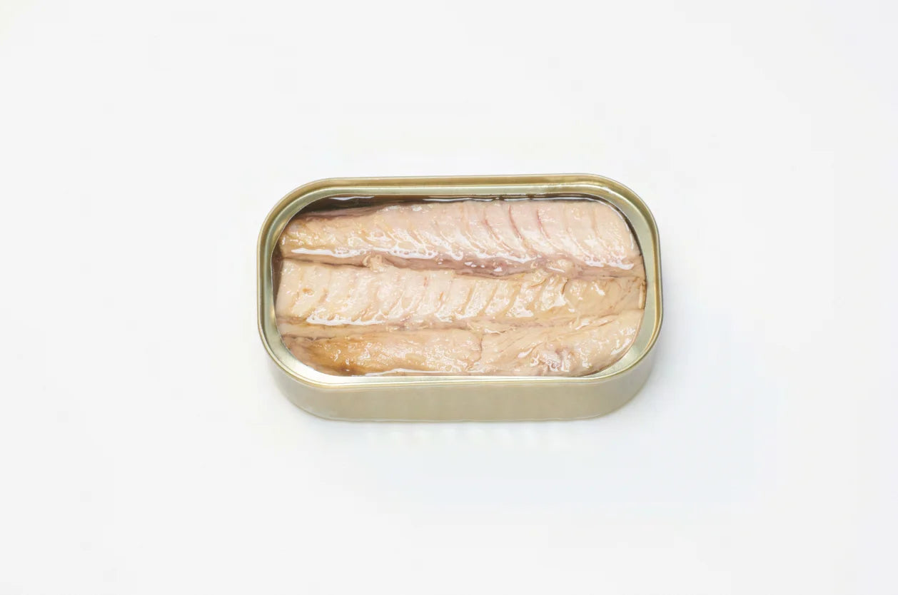A photorealistic close-up of a white tray containing three PYSCIS cans labeled "3-Pack Little Bullet Tuna Filets" (SKU: AUX22). The label features the PYSCIS brand logo and highlights the product's use of sustainable fishing practices. A blue and green gradient adorns the label in the background. The image focuses on a single can tilted slightly forward, revealing a glimpse of the flaky, white tuna meat bathed in olive oil through the transparent window in the can.